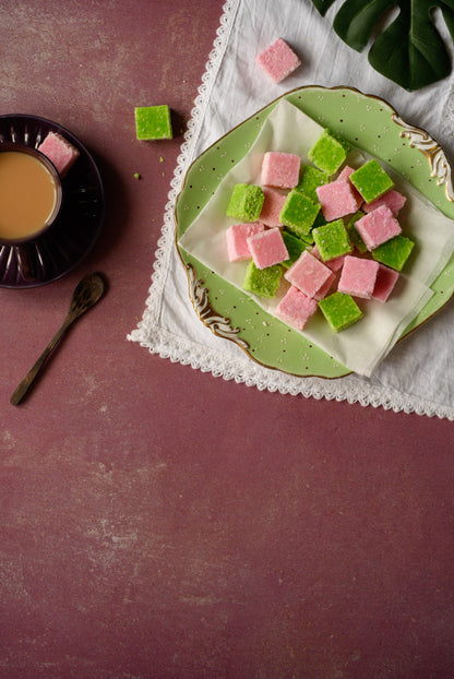 Green and pink square-shaped coconut toffee on a decorative plate with a cup of coffee and spoon on a maroon 