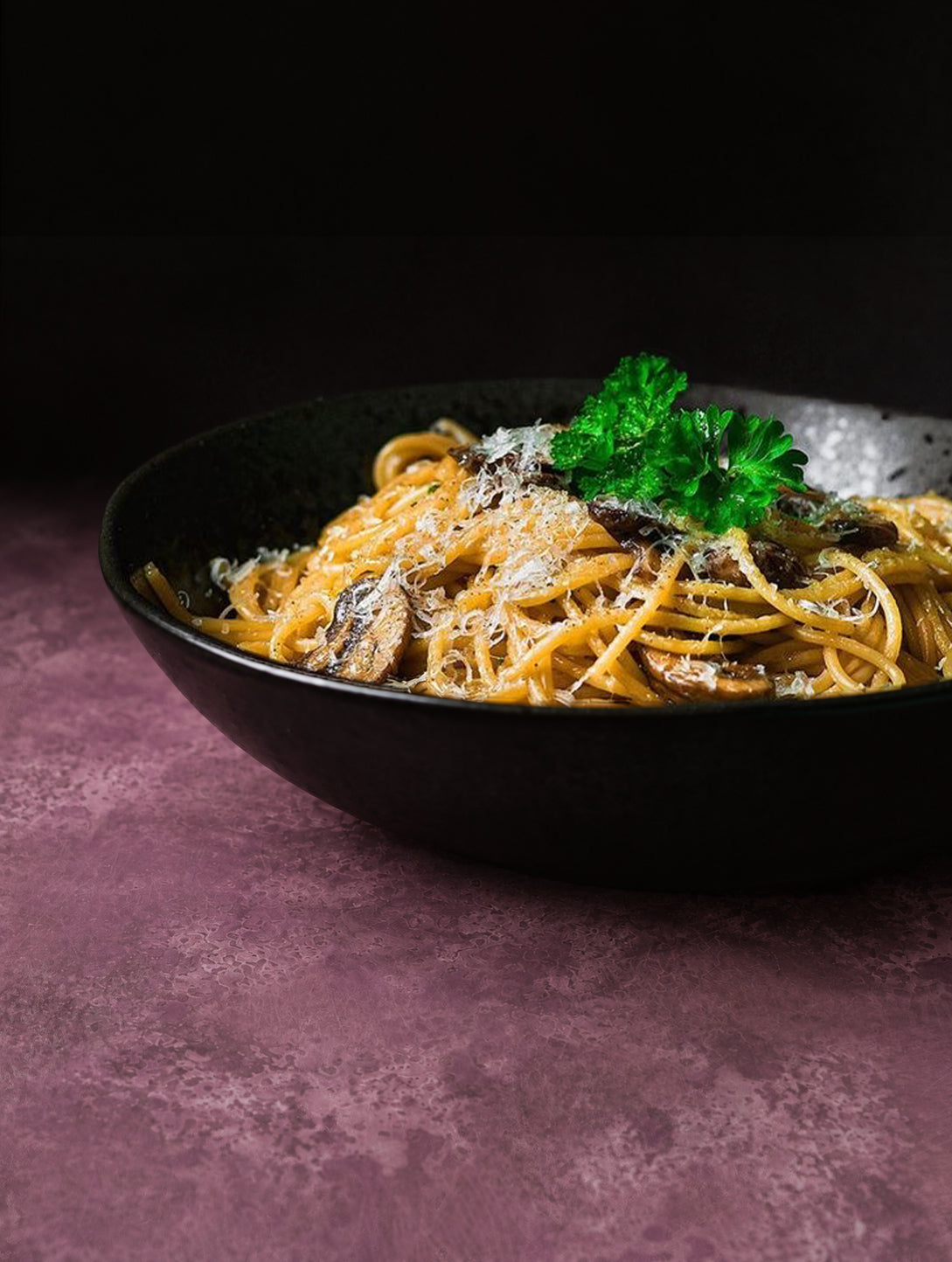 A close-up of a black bowl filled with spaghetti topped with Parmesan and a green parsley garnish, set on a mottled, textured vinyl photography backdrop in dusty mauve-purple tones with lighter and darker patches across the surface.