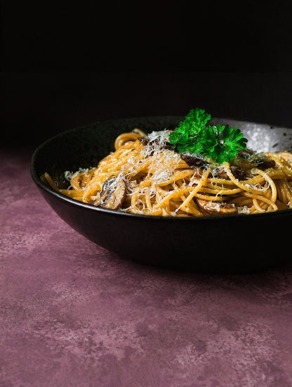 A close-up of a black bowl filled with spaghetti topped with Parmesan and a green parsley garnish, set on a mottled, textured vinyl photography backdrop in dusty mauve-purple tones with lighter and darker patches across the surface.