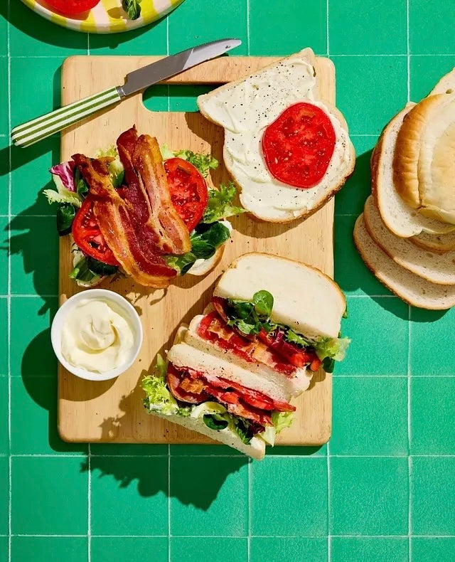 A styled overhead food scene showing two BLT-style sandwiches on a wooden cutting board, with bacon, lettuce, tomato, mayonnaise, bread slices, and a knife on a bright green tiled vinyl photography backdrop.