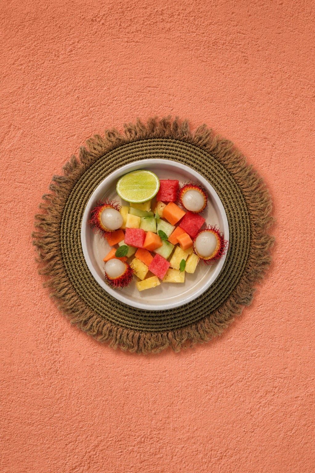 A top-down view of a white bowl filled with pieces of watermelon, mango, pineapple and lychees, garnished with a lime wedge, set on a round woven natural placemat against a warm orange coral stucco-effect vinyl photography backdrop.