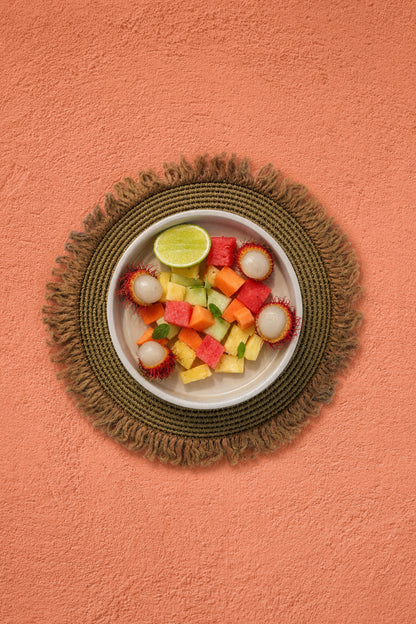 A top-down view of a white bowl filled with pieces of watermelon, mango, pineapple and lychees, garnished with a lime wedge, set on a round woven natural placemat against a warm orange coral stucco-effect vinyl photography backdrop.