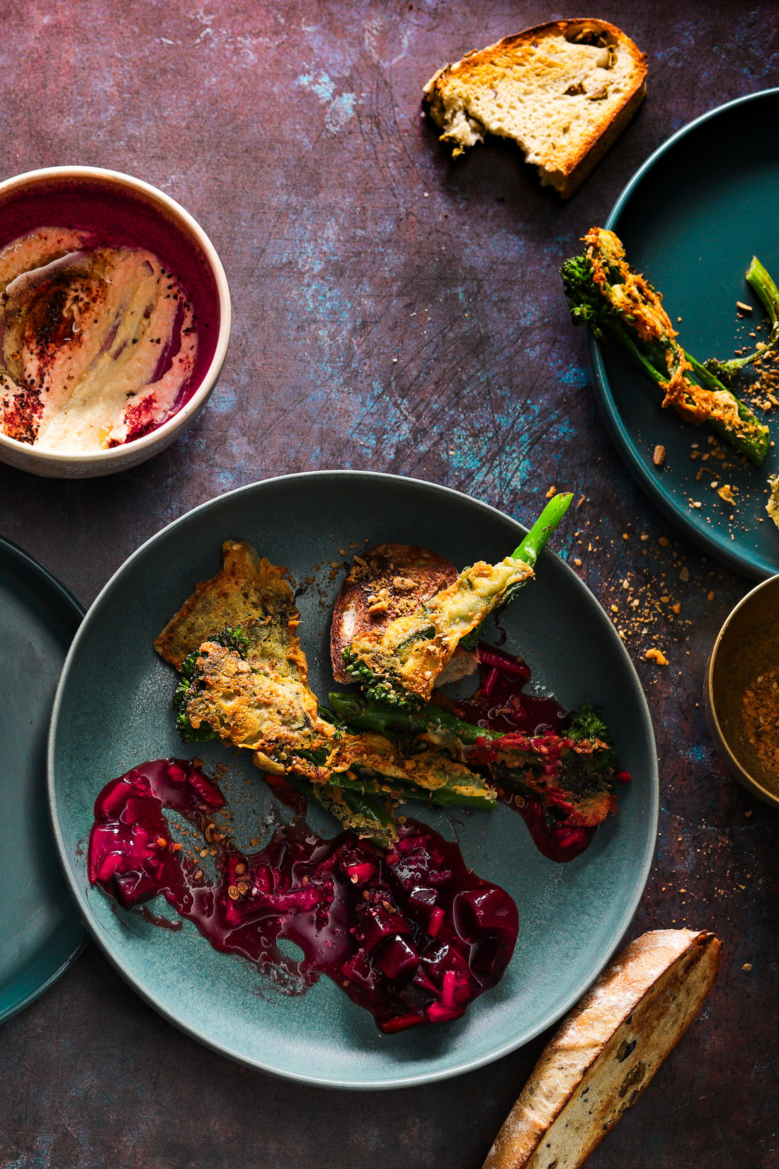 A flat lay of green ceramic plates and nearly finished food styled on a vinyl photography backdrop, with a mottled mix of warm browns, deep plum tones and oxidised blue highlights.