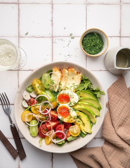Colorful salad with avocado, tomatoes, and onions in a white bowl styled on a white tile effect photography backdrop with a subtle gray grid lines texture.