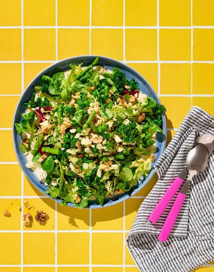 A top-down view of a green salad in a blue bowl, set on a yellow tile effect vinyl photography backdrop with white grout lines, next to a grey-and-white striped napkin with pink-plastic handled utensils.