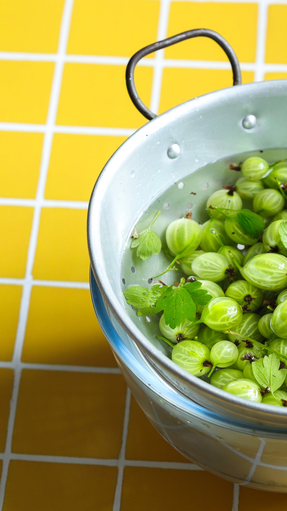 Colander of fresh gooseberries on a yellow tile effect vinyl photography backdrop with white grout lines.