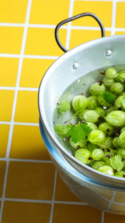 Colander of fresh gooseberries on a yellow tile effect vinyl photography backdrop with white grout lines.