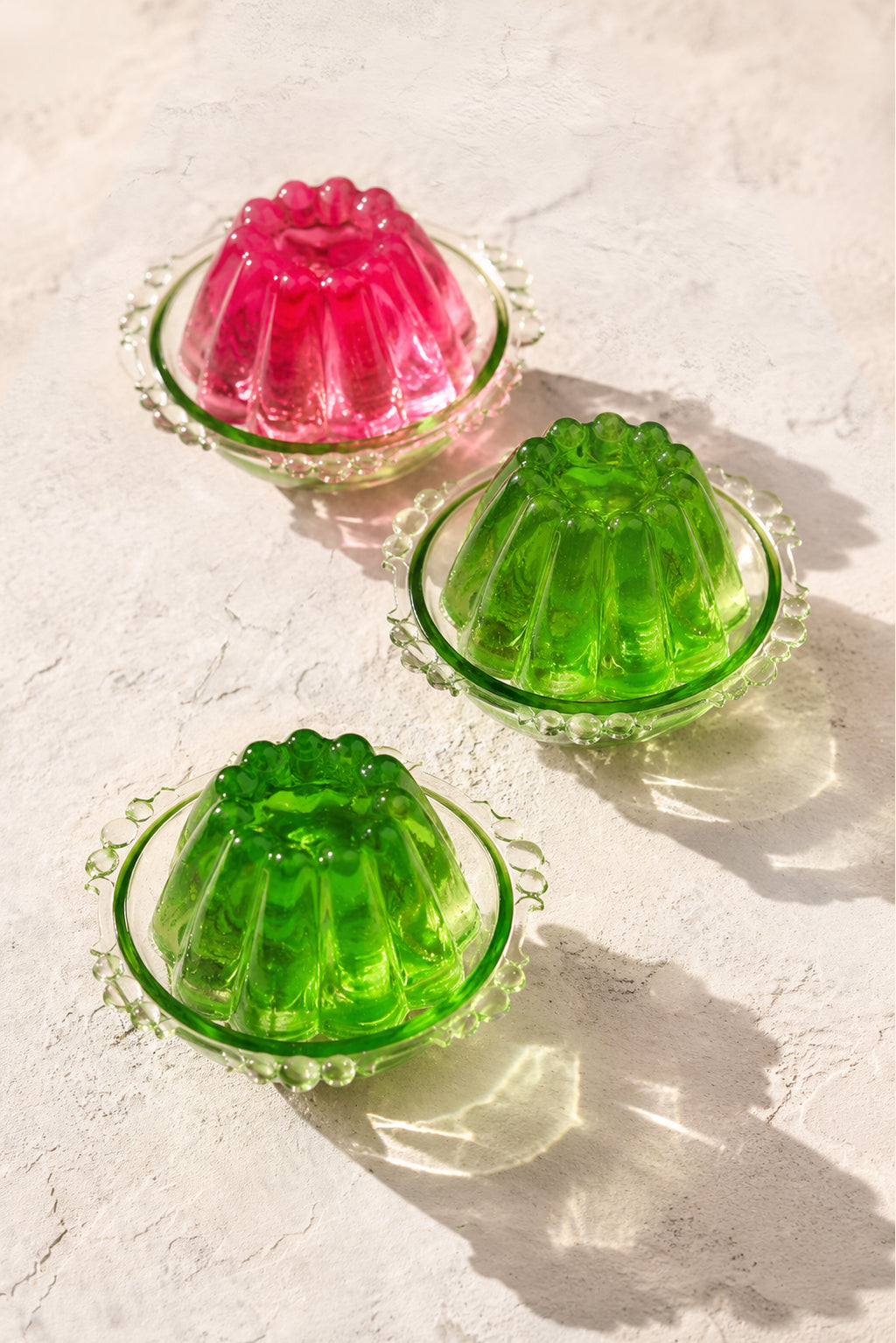 A styled image of pink and green mini jellies in vintage green glass dishes against a white vinyl photography backdrop with a plastered wall effect.