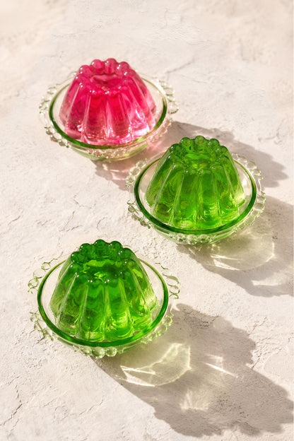 A styled image of pink and green mini jellies in vintage green glass dishes against a white vinyl photography backdrop with a plastered wall effect.
