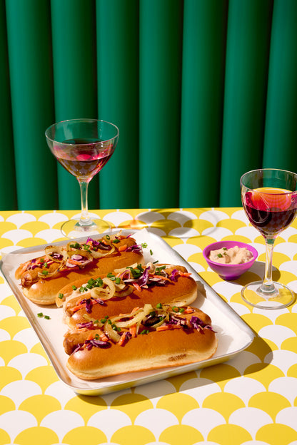 A brightly lit tray of three hot dogs and two glasses of red wine, styled on a scalloped yellow and white tile effect graphic photography backdrop.