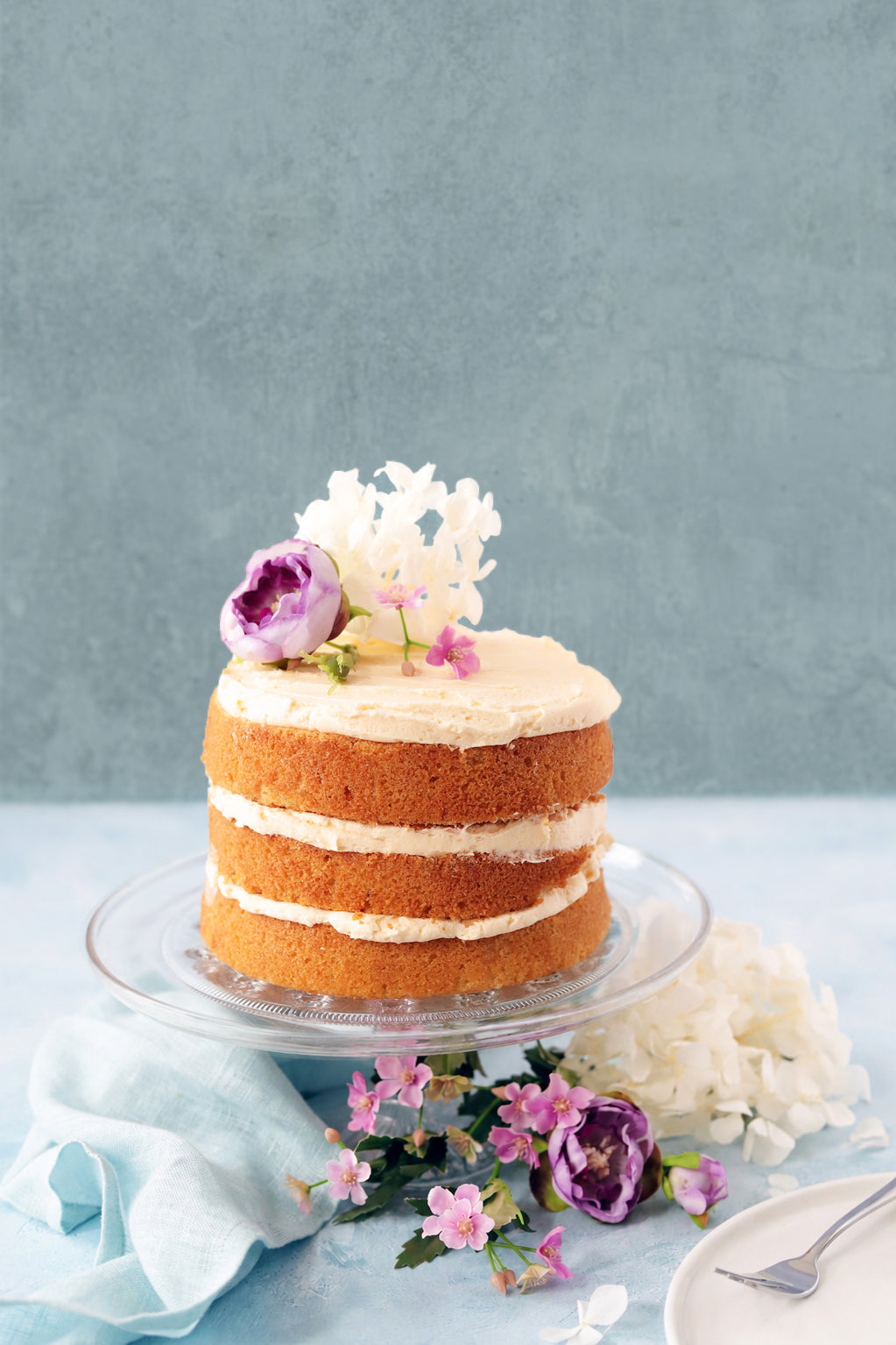 Naked cake with white frosting and floral decorations shot against a light blue and grey food photography backdrop