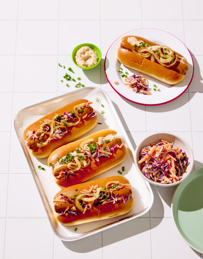 Three hot dogs with toppings on a white tray, accompanied by small bowls of condiments styled on a white tile-effect vinyl photography backdrop with grid lines.