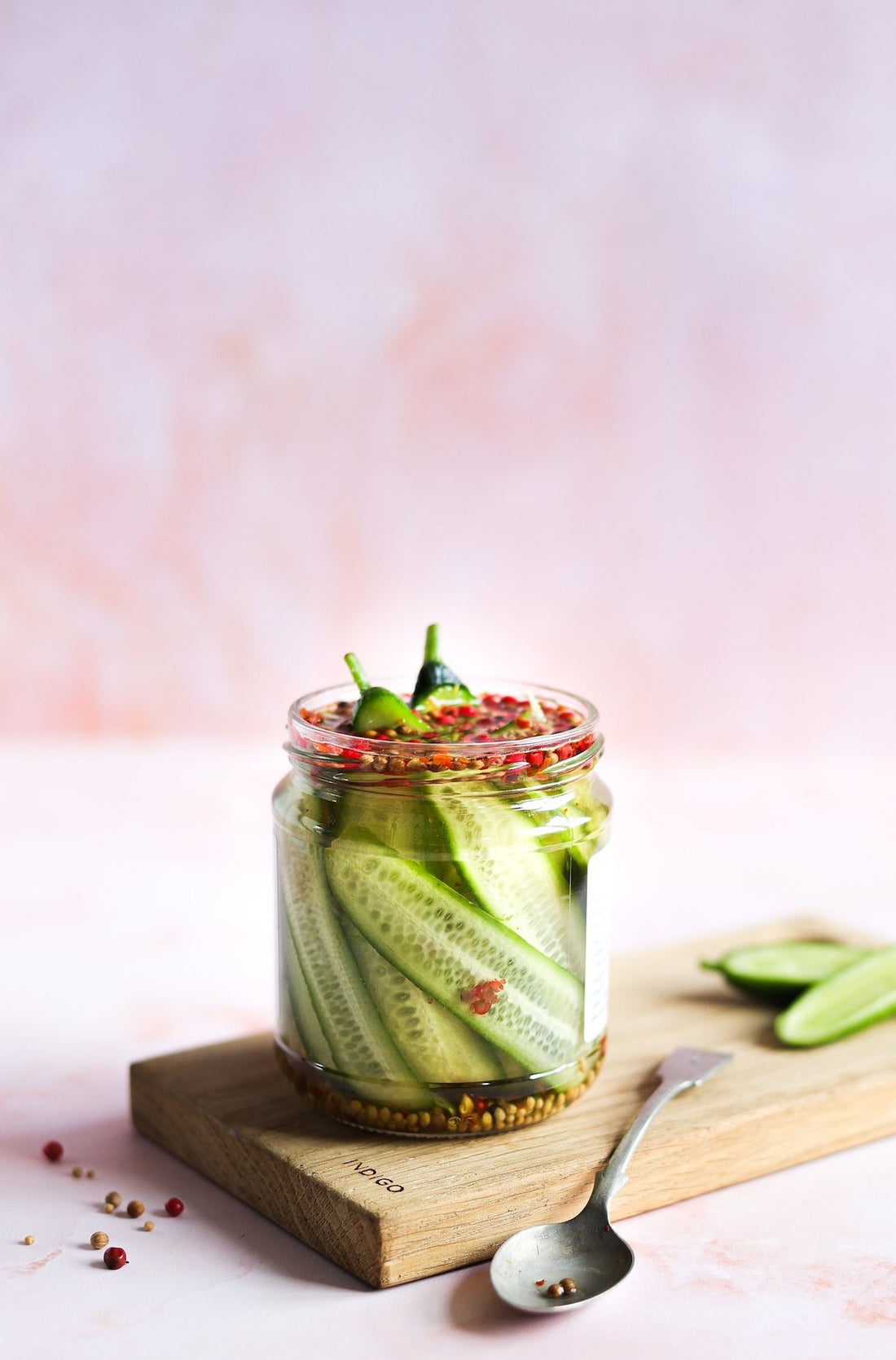 A glass jar of pickled cucumbers styled against a hand-painted effect photography backdrop in light peach pink with water marked textures.