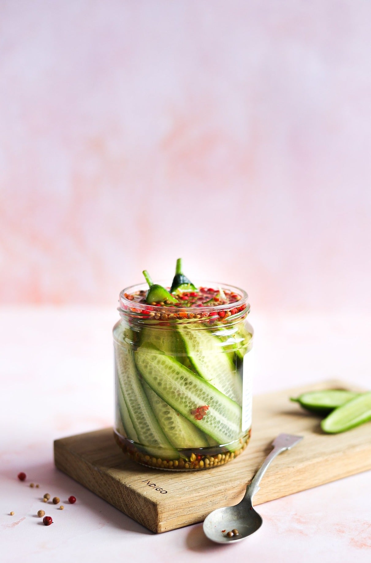A glass jar of pickled cucumbers styled against a hand-painted effect photography backdrop in light peach pink with water marked textures.