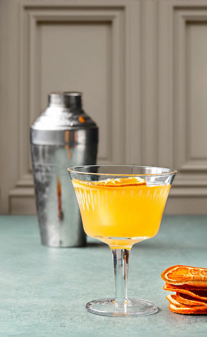 Orange cocktail in a glass with a shaker and orange slices on a light blue surface.
