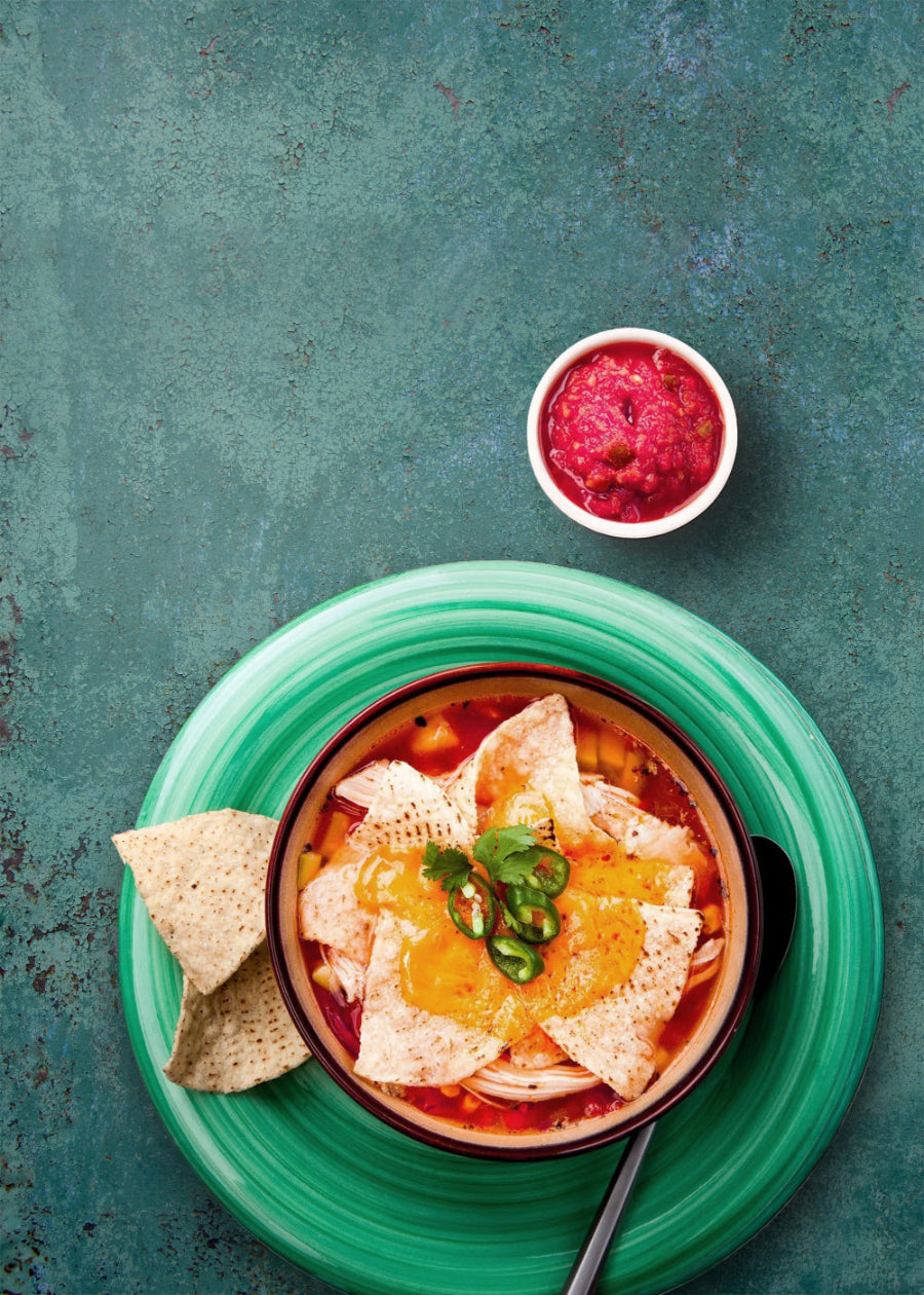 Bowl of soup with tortilla chips and a side of salsa on a green plate against a teal vinyl photography backdrop with a rusted metal details