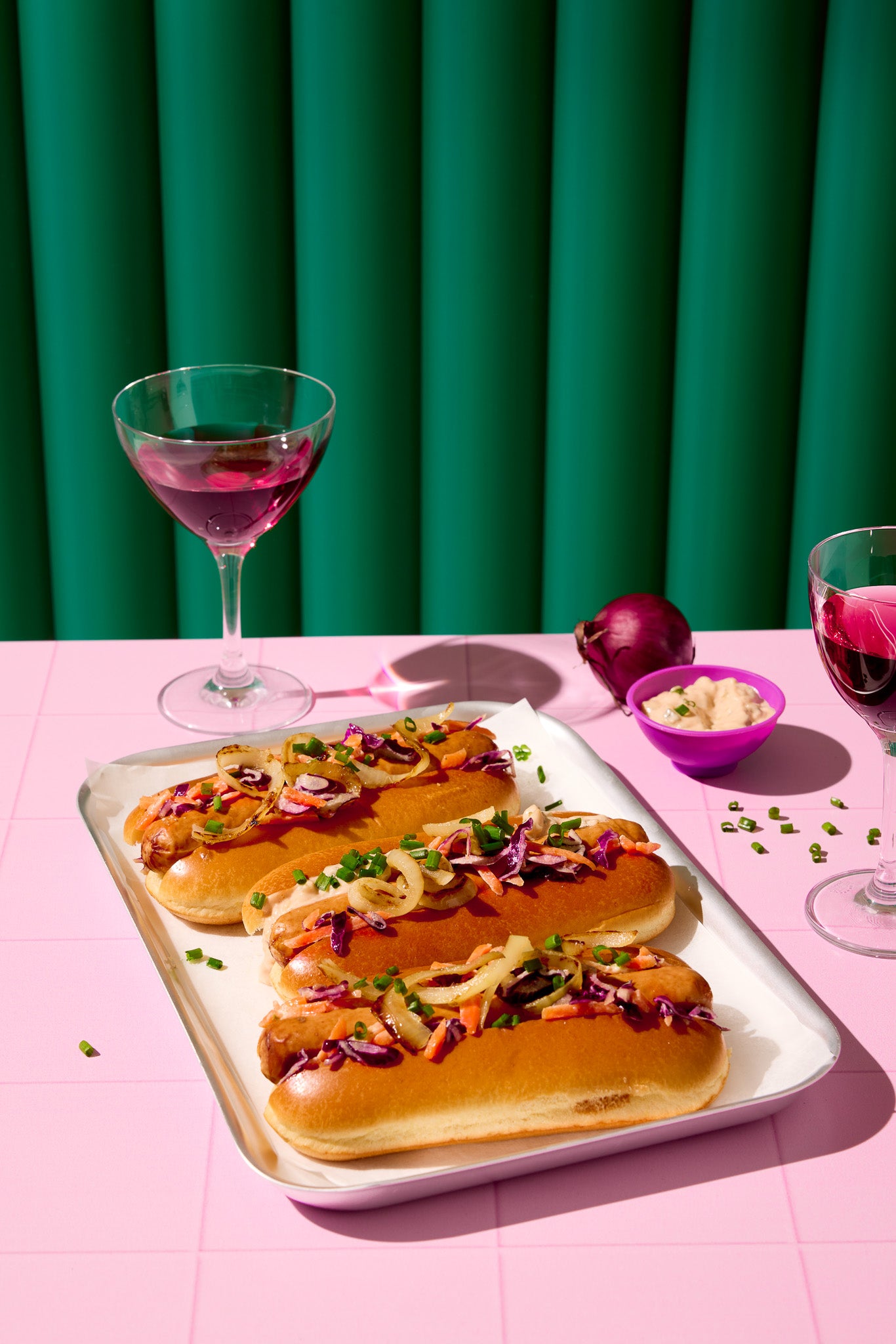 A commercial food photo of a tray of hotdogs with red wine glasses styled against an emerald green ribbed vinyl photography backdrop with a 3D effect.