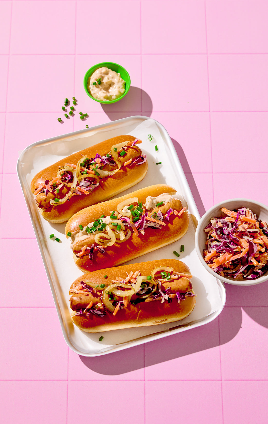Three hot dogs with toppings on a white tray against a pink tiled background