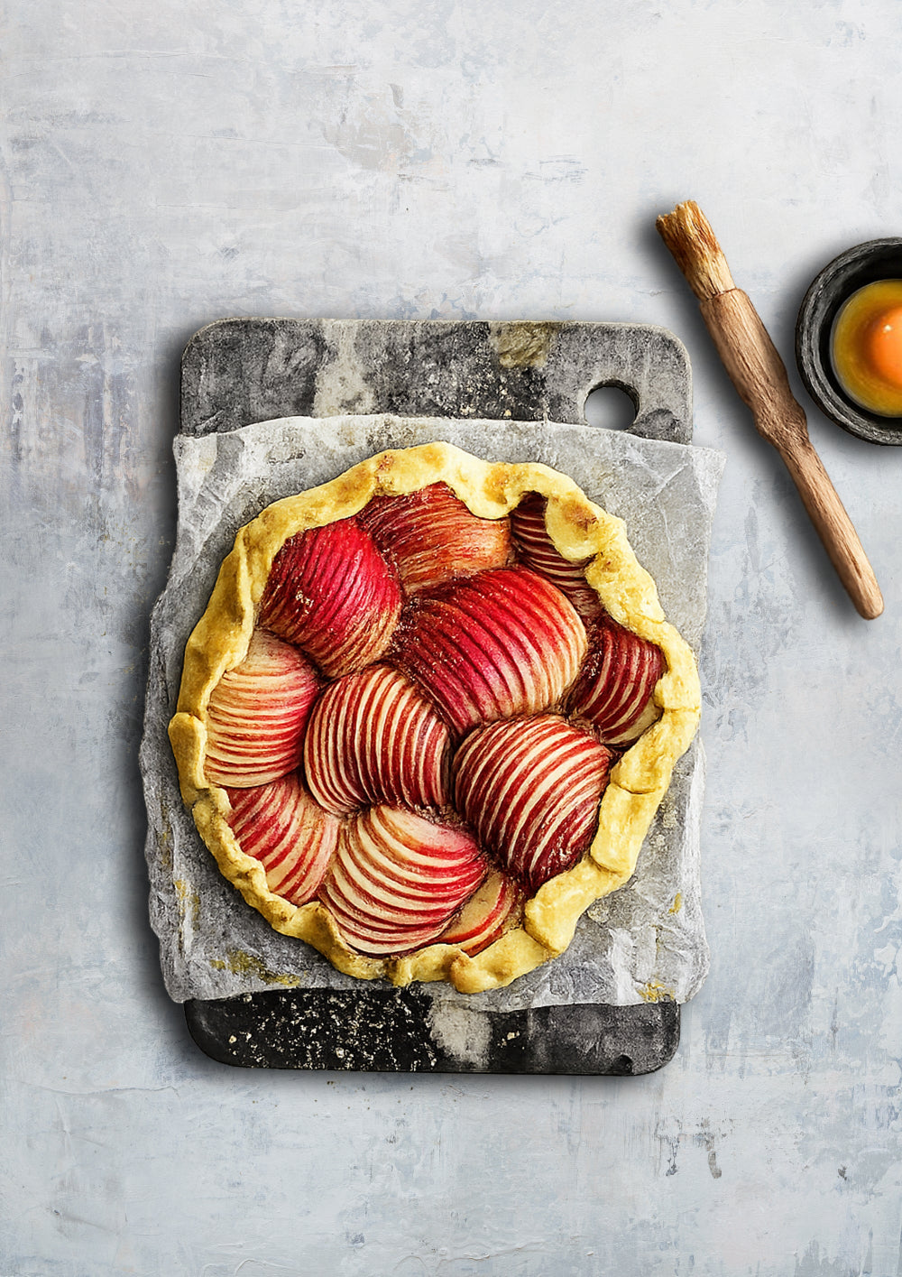 Apple pie on a stone cutting board with a wooden rolling pin and a bowl of flour in the background.
