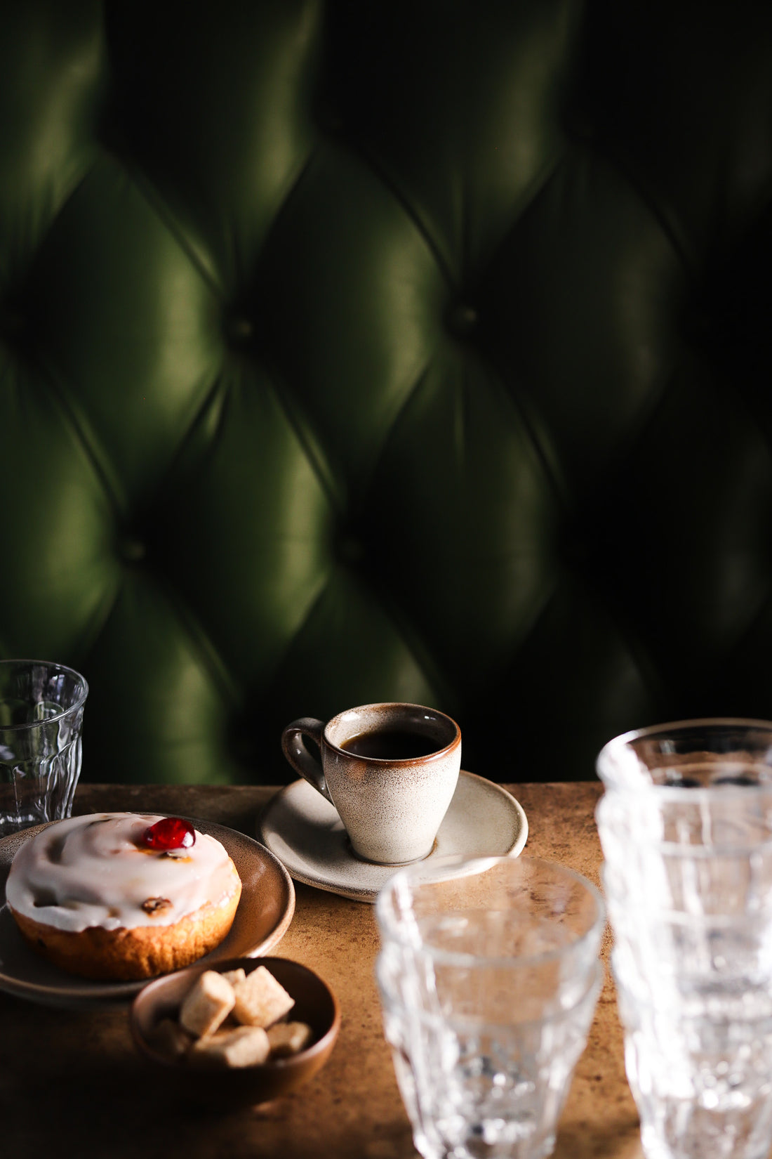 A cup of coffee and pastry against a green leather effect photography backdrop with button details, resembling a Chesterfield sofa.