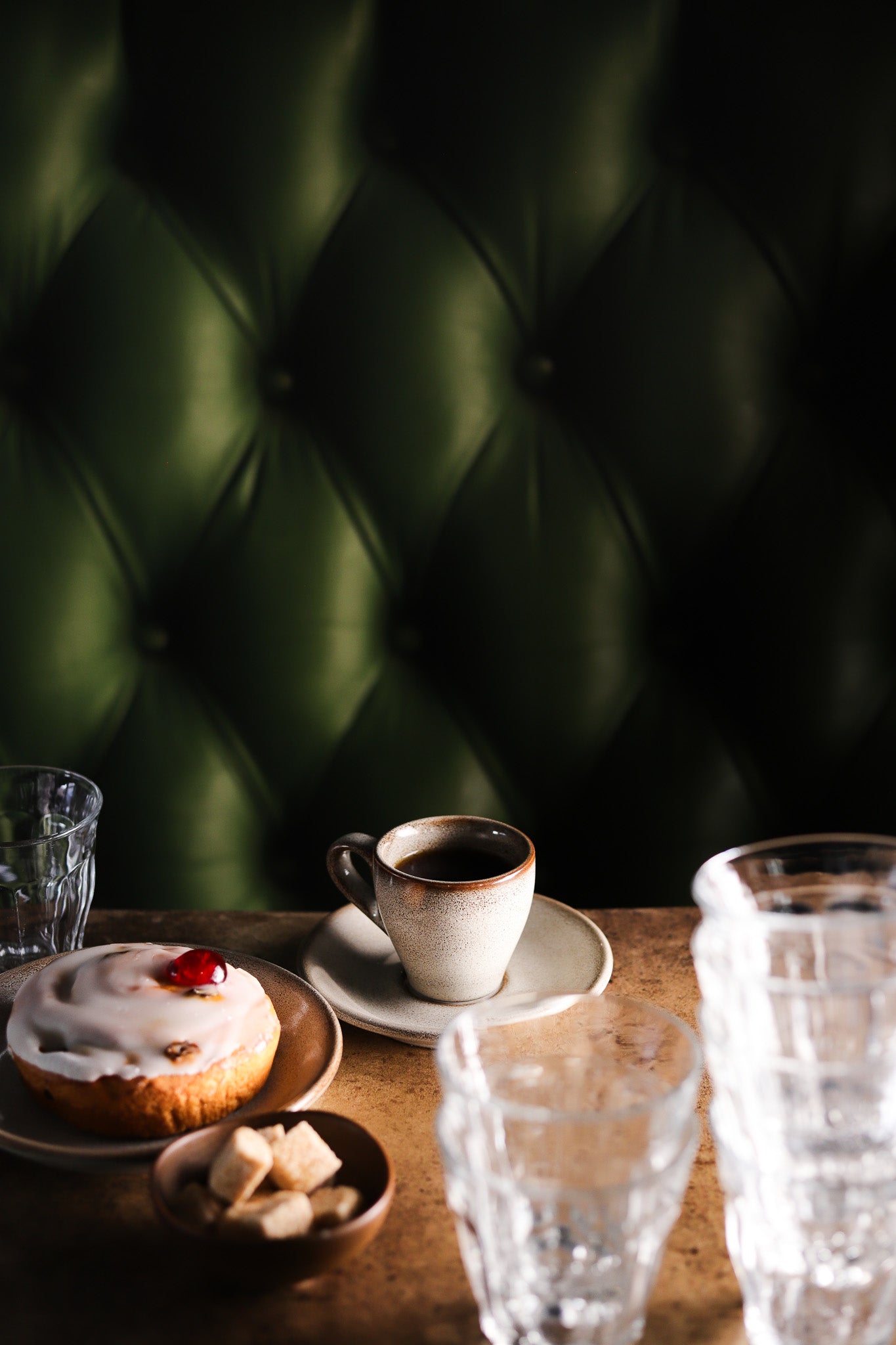 A cup of coffee and pastry against a green leather effect photography backdrop with button details, resembling a Chesterfield sofa.