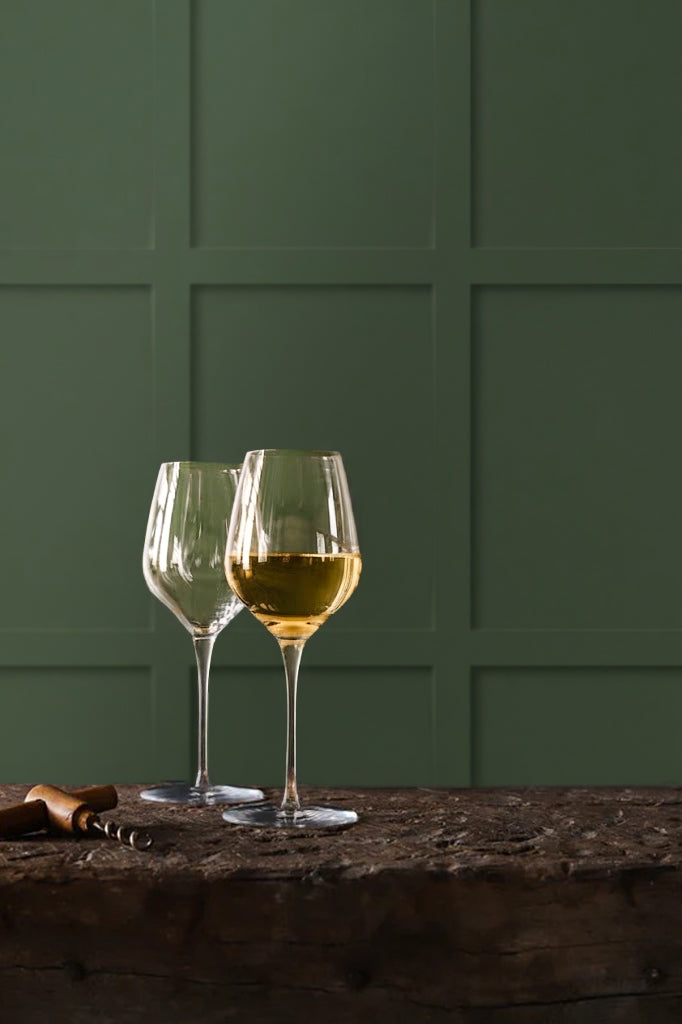 Two clear wine glasses on a rustic wooden table, with one glass partially filled with white wine and the other empty, set against an olive green wood paneled wall effect photography backdrop for food and product styling.