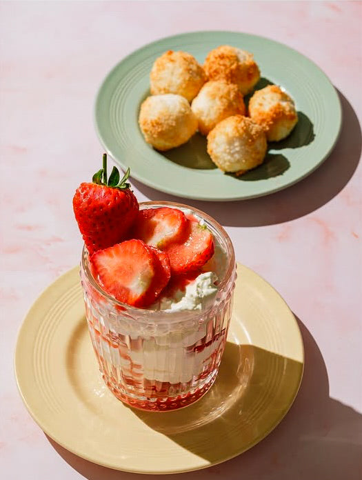 A dessert glass topped with fresh strawberries and cream on a yellow plate with a green plate of coconut macroons shot on a pink marble photography backdrop.