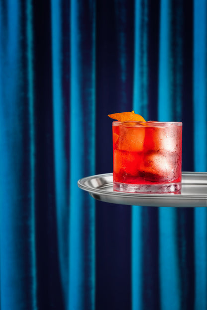 Red cocktail with an orange twist on a silver tray against a peacock blue silk velvet curtain effect vinyl photography backdrop.