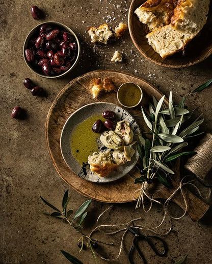 An overhead styled food scene on a warm mottled brown vinyl photography backdrop featuring a plate with bread, artichoke, olives, olive oil, a small bowl of dressing, and a bunch of olive branches tied with twine. 