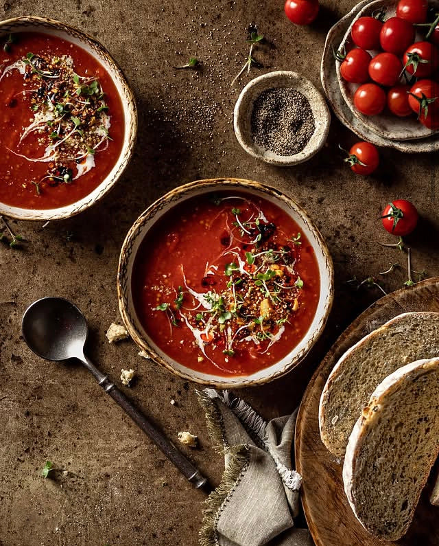 Two rustic bowls of tomato soup garnished with microgreens, styled on a warm brown mottled vinyl photography backdrop alongside sliced bread and cherry tomatoes.