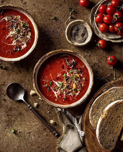 Two rustic bowls of tomato soup garnished with microgreens, styled on a warm brown mottled vinyl photography backdrop alongside sliced bread and cherry tomatoes.