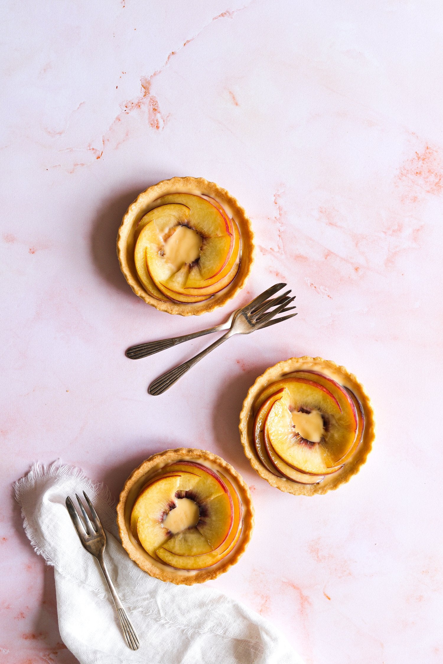 Three small fruit tartlets with sliced peaches styled on a soft peach marble photography backdrop with organic swirling patterns.