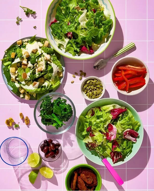 An overhead flat-lay of multiple bowls of salad arranged on a pink tile-effect vinyl photography backdrop. The salads include green lettuce, broccoli, radicchio, red peppers, olives, walnuts, and dressing, with serving utensils and garnishes placed around them.