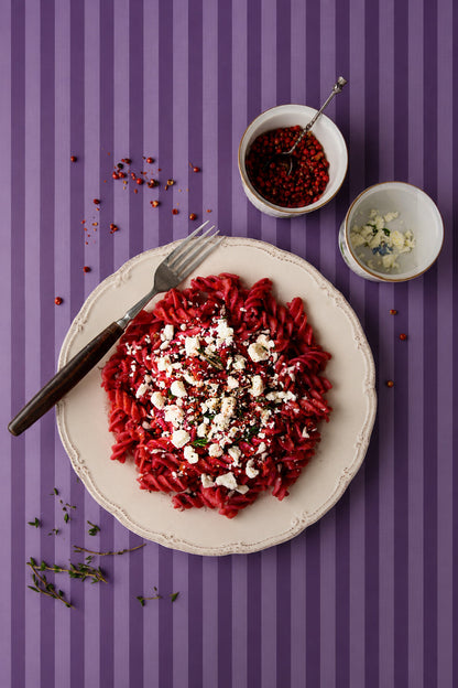 Top down image of a vintage plate of beetroot fusilli pasta styled on a purple and lavender striped vinyl photography backdrop.