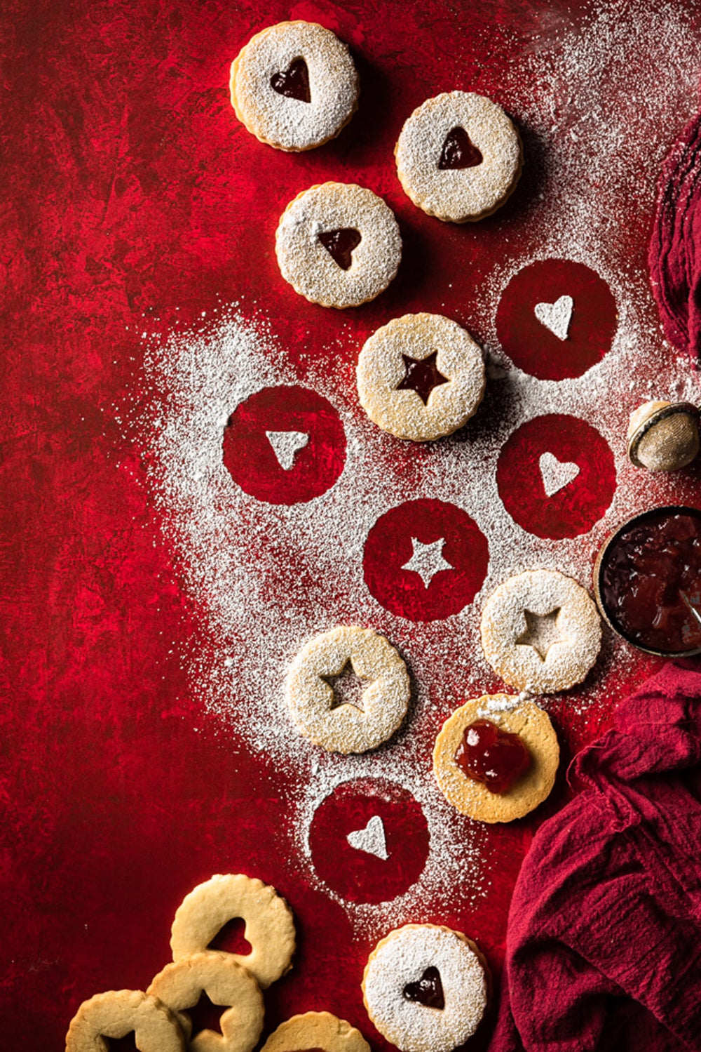 Flat lay of Linzer cookies with jam on a red plaster-effect photography backdrop with a textured surface.