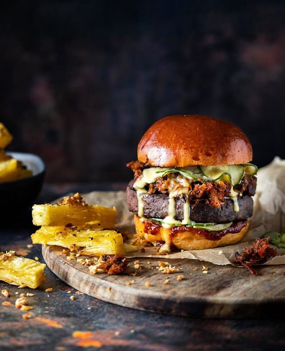 A gourmet burger with multiple layers of meat, melted cheese, and toppings, served on a wooden board with golden fries on a dark brown vinyl photography backdrop.