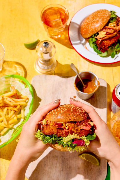 Brighly lit styled burger table scene shot on a saffron yellow wood-effect vinyl photography backdrop with visible wood grain.