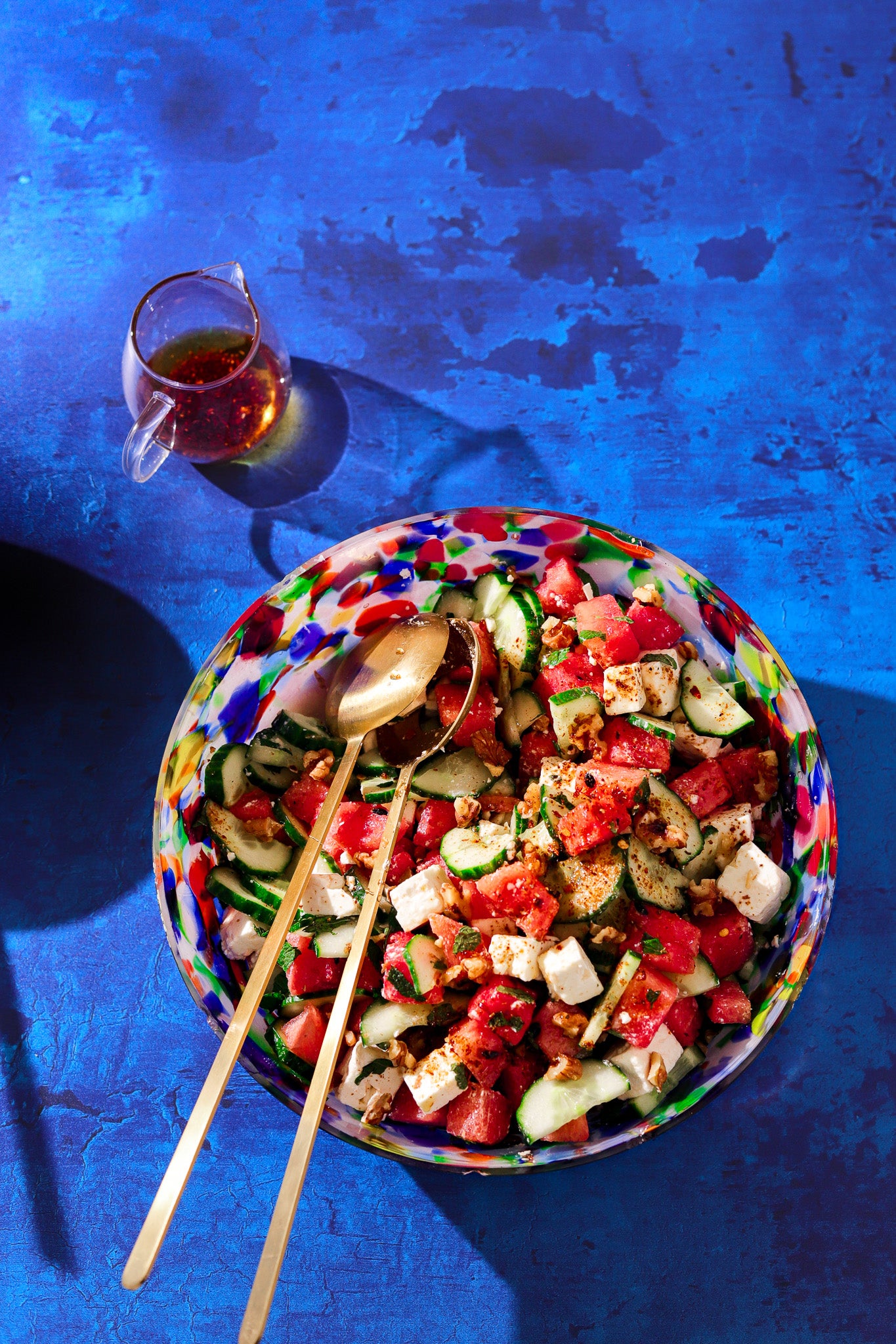 A bowl of watermelon salad in a bright multicoloured glass bowl styled on a cobalt blue vinyl photography backdrop with a layered paint effect over wood.