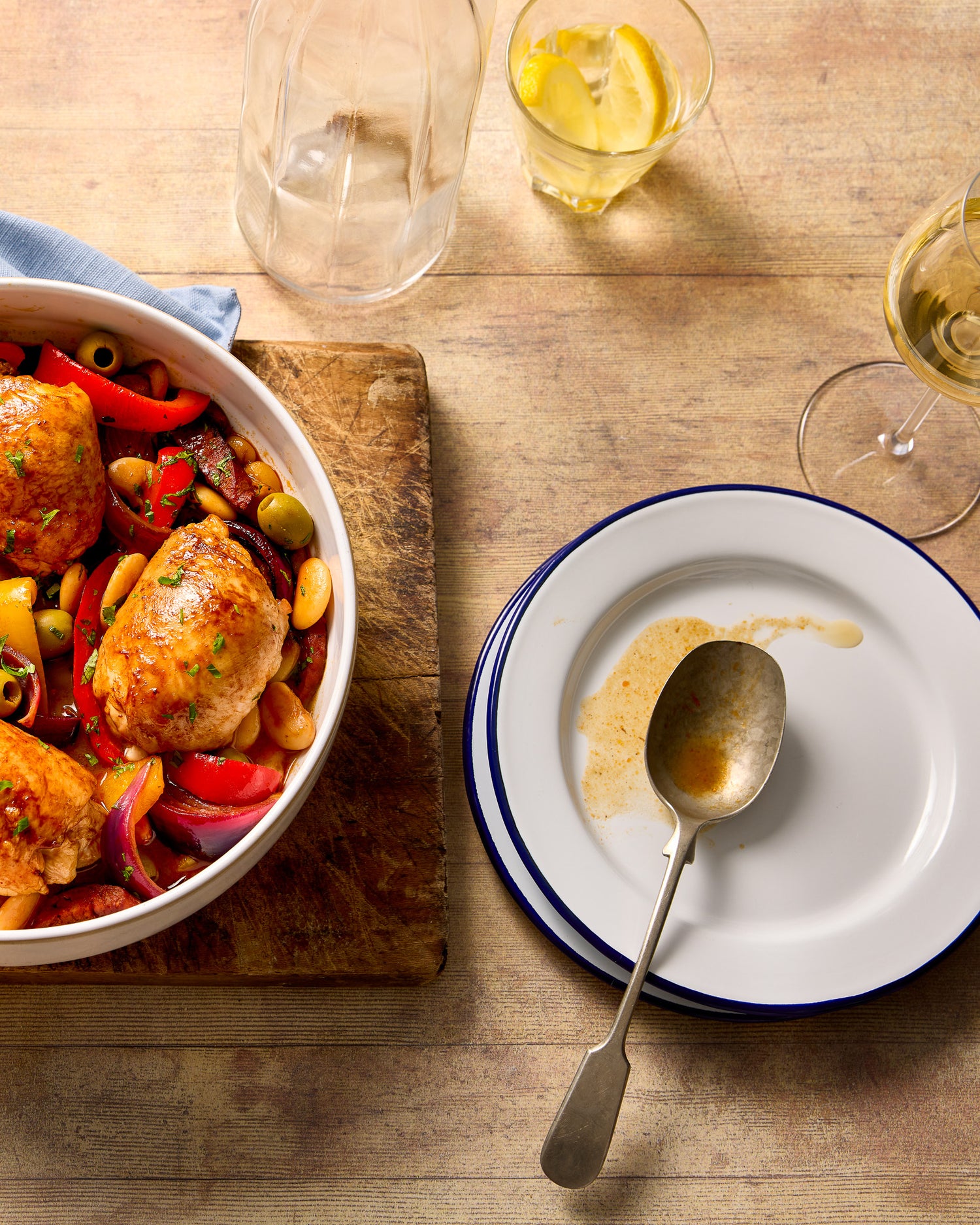 A flat lay of a chicken casserole, glasses and plates styled on a wooden table effect photography backdrop with a washed out pine or oak appearance.