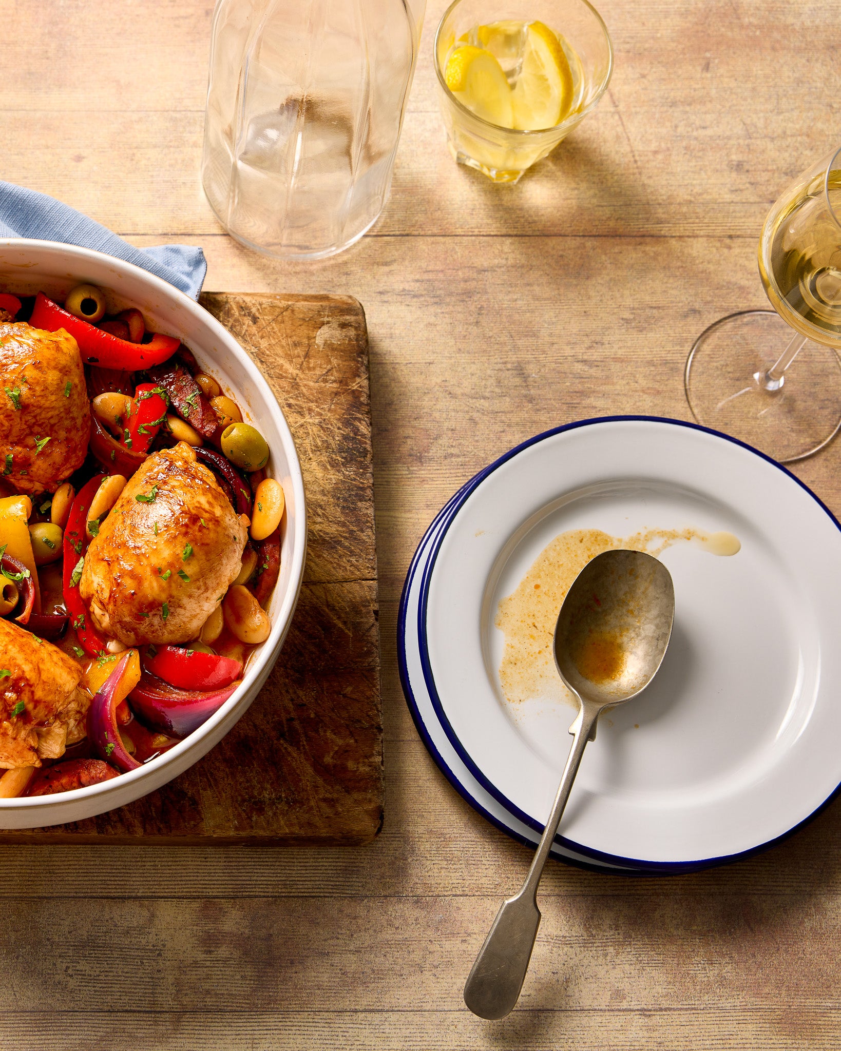 A flat lay of a chicken casserole, glasses and plates styled on a wooden table effect photography backdrop with a washed out pine or oak appearance.
