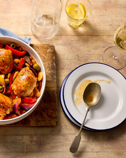 A flat lay of a chicken casserole, glasses and plates styled on a wooden table effect photography backdrop with a washed out pine or oak appearance.