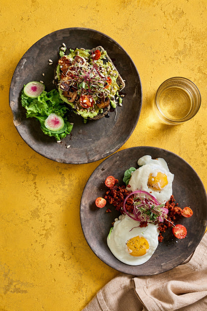 Two rustic dark grey plates of brunch food. The top plate features avocado toast served with lettuce and radishes while the bottom plate features poached eggs and cherry tomatoes. Styled against a yellow vinyl photography backdrop with a weathered plaster design for food styling and product photography.