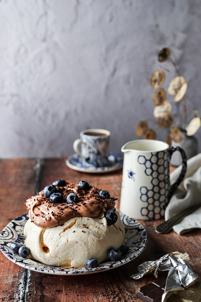A white meringue with chocolate frosting and fresh blueberries on a patterned plate, set on a rustic wooden photography backdrop with a plaster effect backdrop behind.