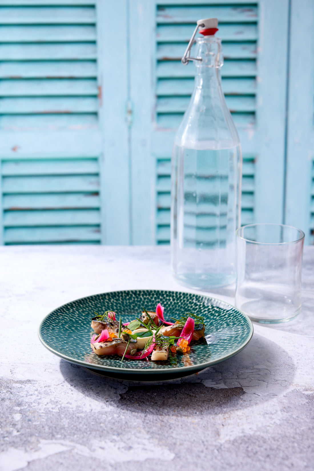 Blue decorative plate and colourful salad with a bottle of water and glass shot against a vinyl photography backdrop featuring a pair of blue wooden shutters with a blurred effect.