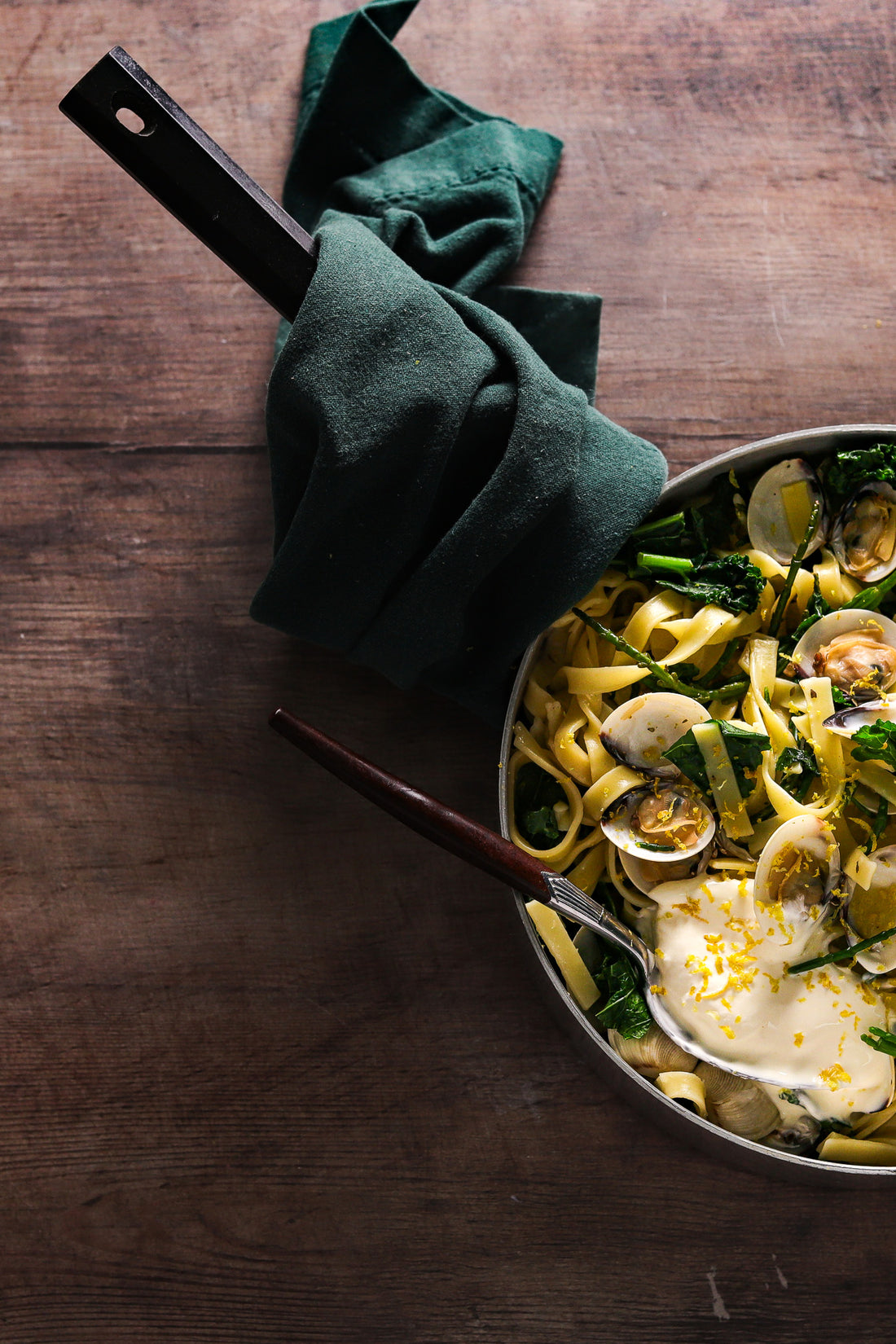 A pan of clam pasta styled on a dark wood table effect photography backdrop with a soft woodgrain effect.