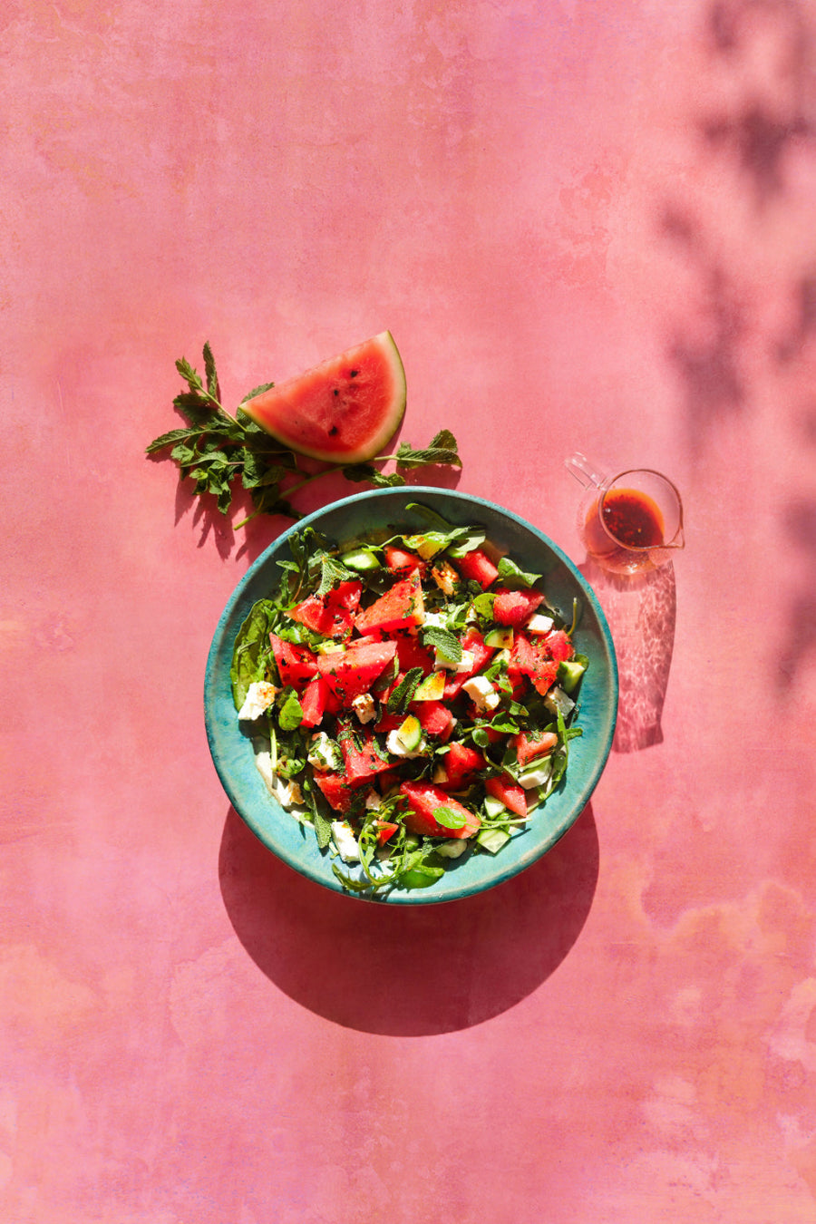 A top-down view of a green bowl filled with a fresh watermelon and mint summer salad, set on a pink and orange vinyl photography backdrop with a mottled watercolour effect.