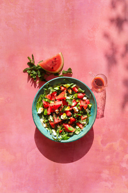 A top-down view of a green bowl filled with a fresh watermelon and mint summer salad, set on a pink and orange vinyl photography backdrop with a mottled watercolour effect.