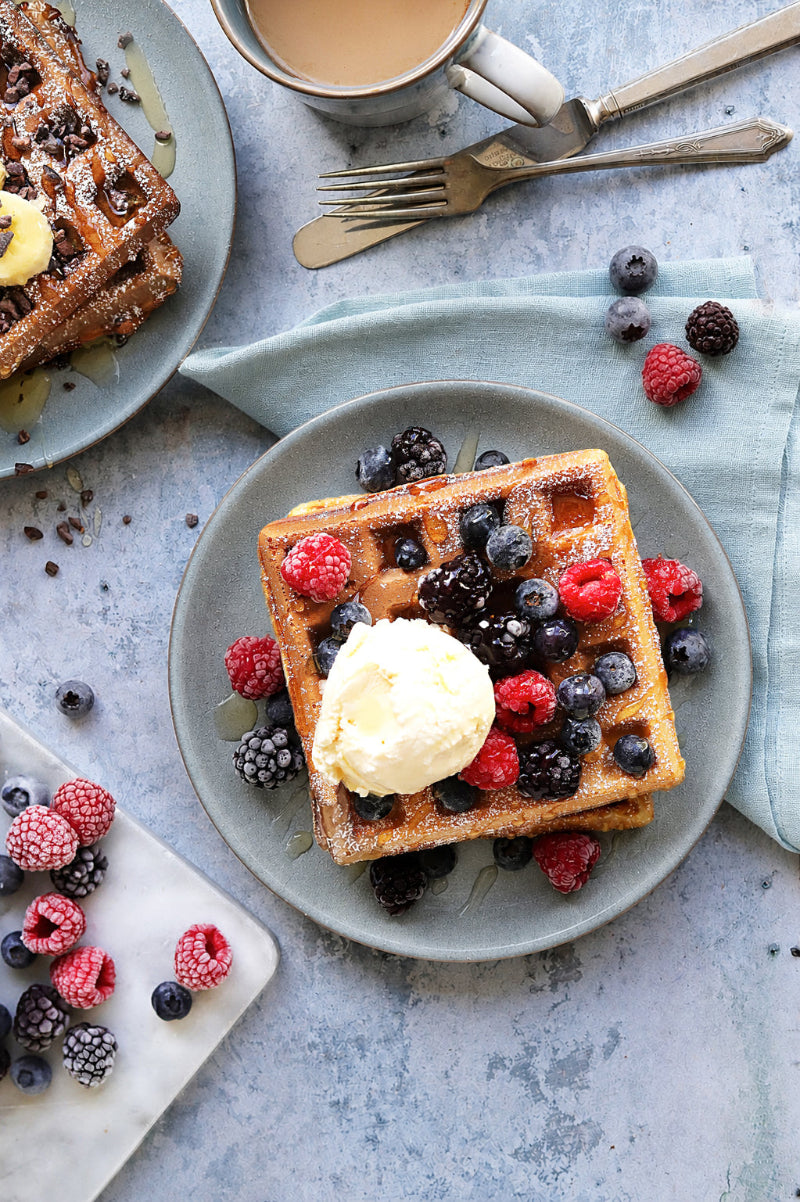 A top-down image of a plate of waffles topped with a scoop of vanilla ice cream and fresh berries, with additional plates, utensils, and a cup of coffee styled on a pale blue waterproof vinyl photography backdrop.