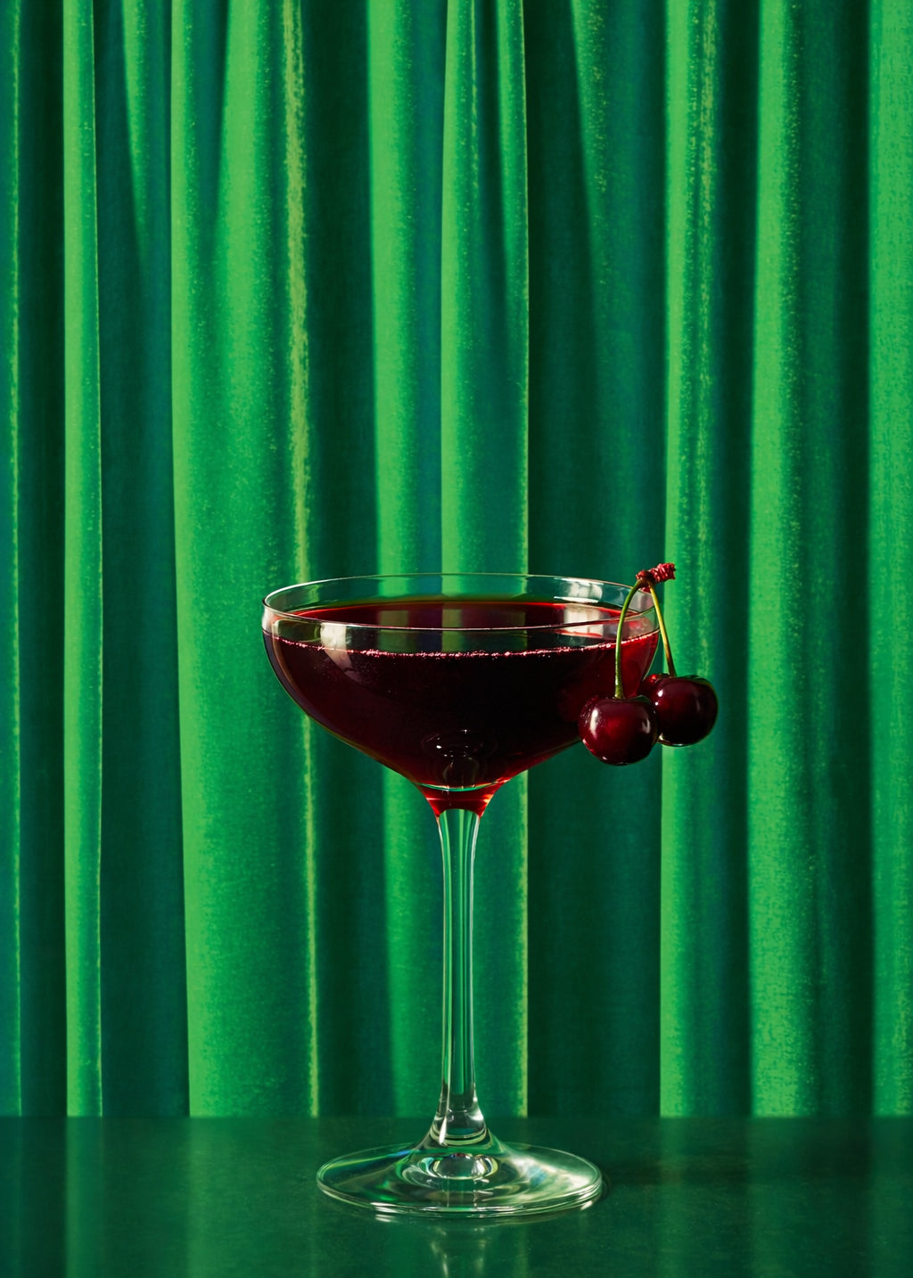 Cherry Manhattan cocktail with fresh red cherries in a coupe glass against a grass green silk curtain-style vinyl photography backdrop.