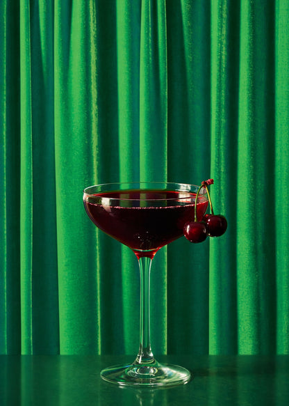 Cherry Manhattan cocktail with fresh red cherries in a coupe glass against a grass green silk curtain-style vinyl photography backdrop.